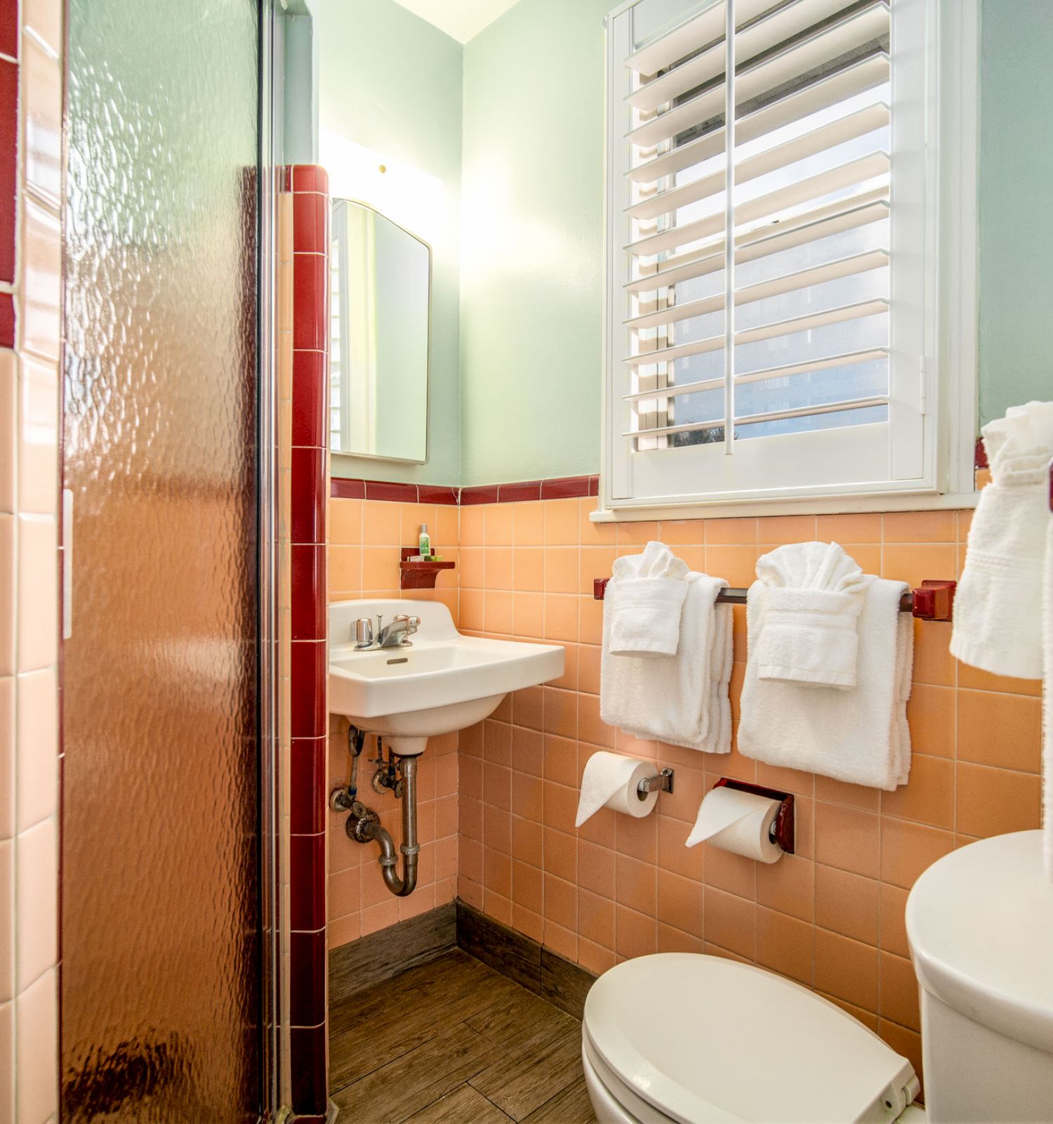 The image depicts a small bathroom with pastel-colored tiles, a walk-in shower, a small sink, a toilet, and neatly arranged towels on racks.