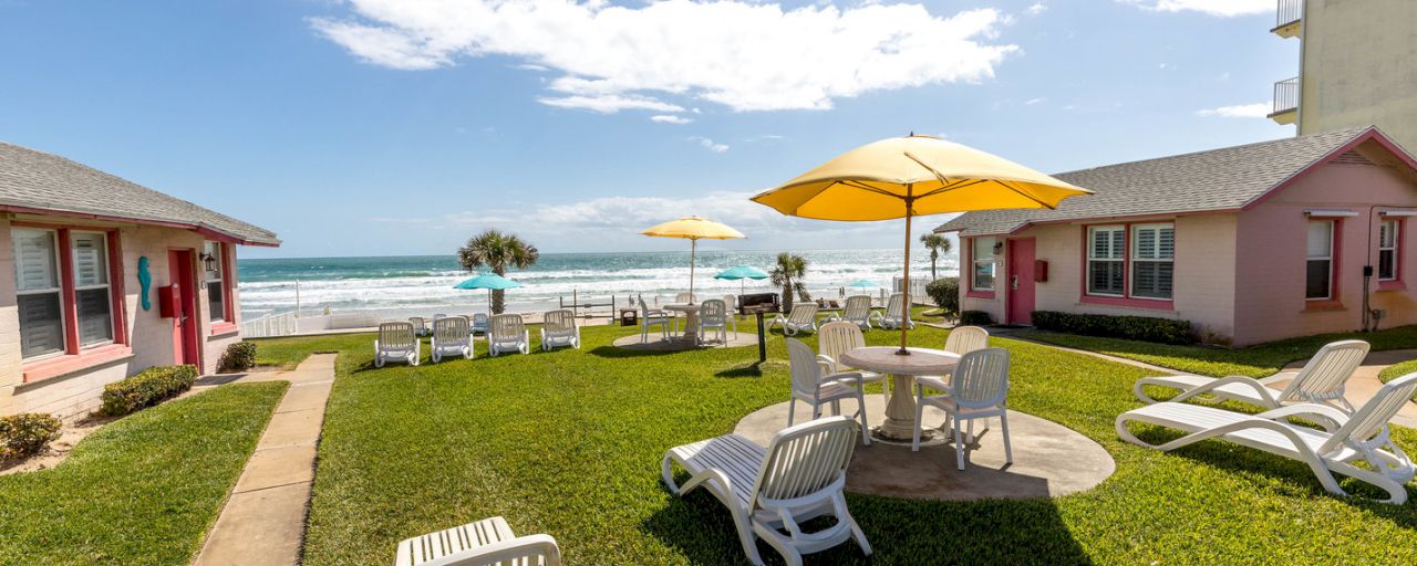 A beachside resort with pink buildings, a grassy area with white furniture and yellow umbrellas, and a view of the ocean.