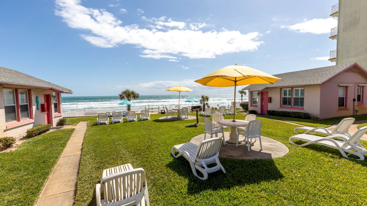 A beachfront resort with pink buildings, a grassy area with white furniture, and a view of the ocean.
