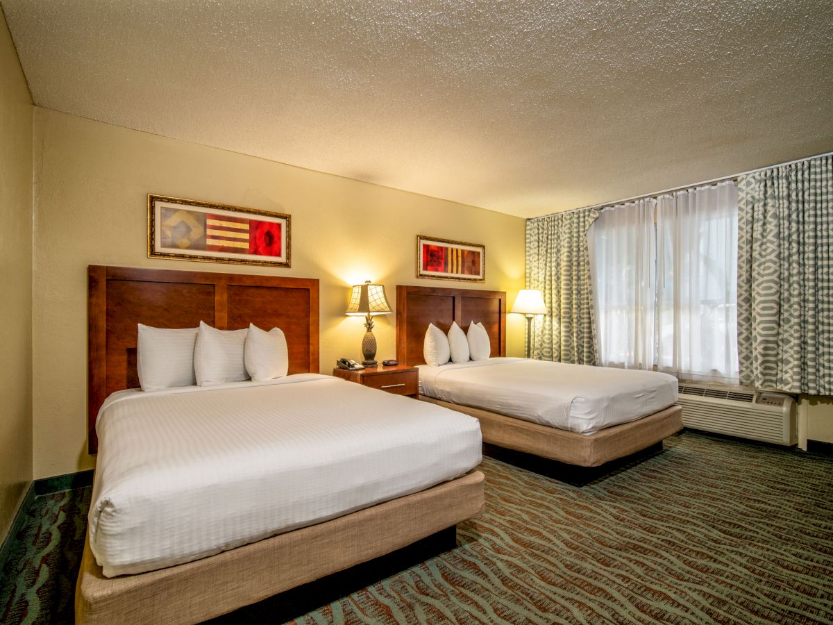 A hotel room with two double beds, artwork above them, a nightstand with lamps, and a window with patterned curtains to the right.