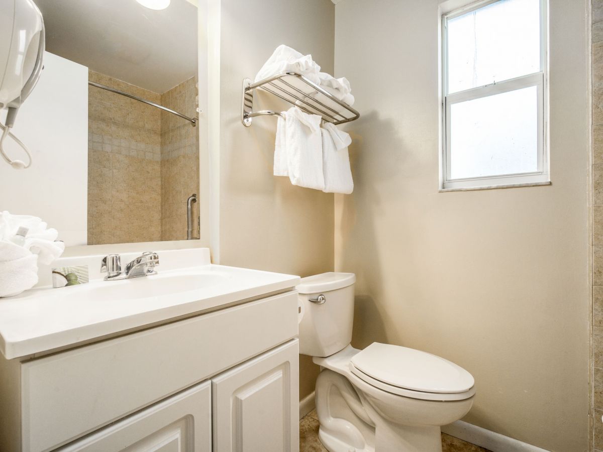 The image shows a clean, simple bathroom with a white sink, toilet, towel rack with towels, window, and mirror, all in a beige-toned setting.