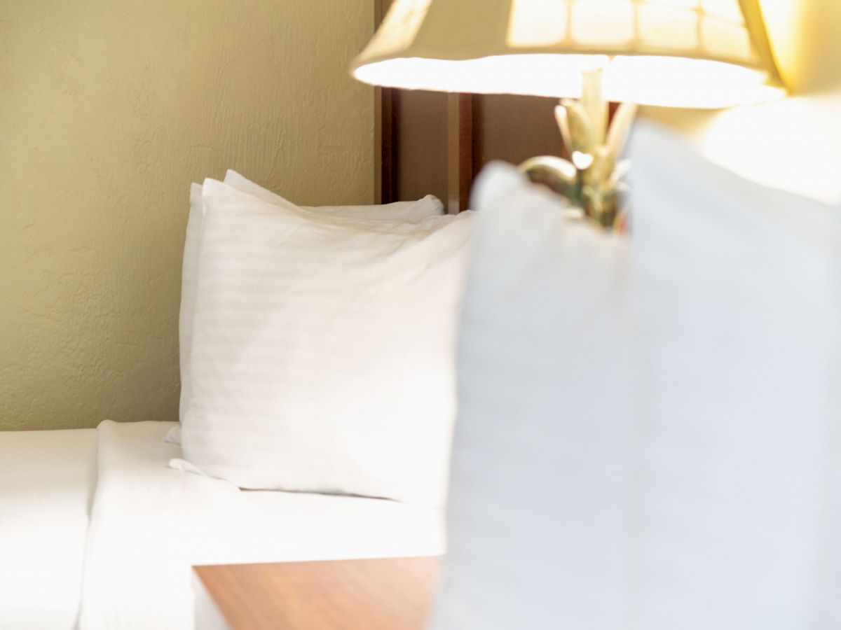 The image shows a cozy bedroom with a bed, two white pillows, and a lamp on a bedside table, creating a warm and inviting atmosphere.