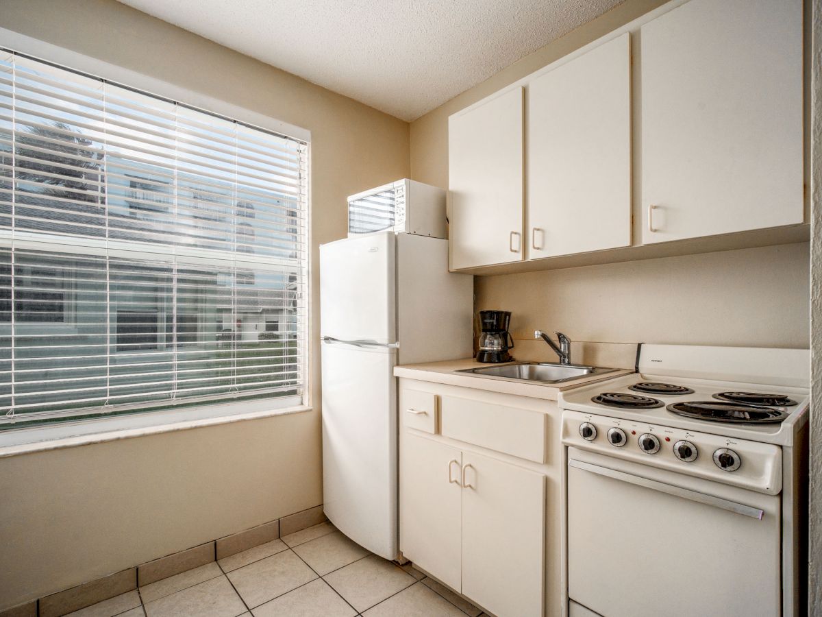 A small kitchen with a window, fridge, microwave, coffee maker, sink, cabinets, and stove with four burners.