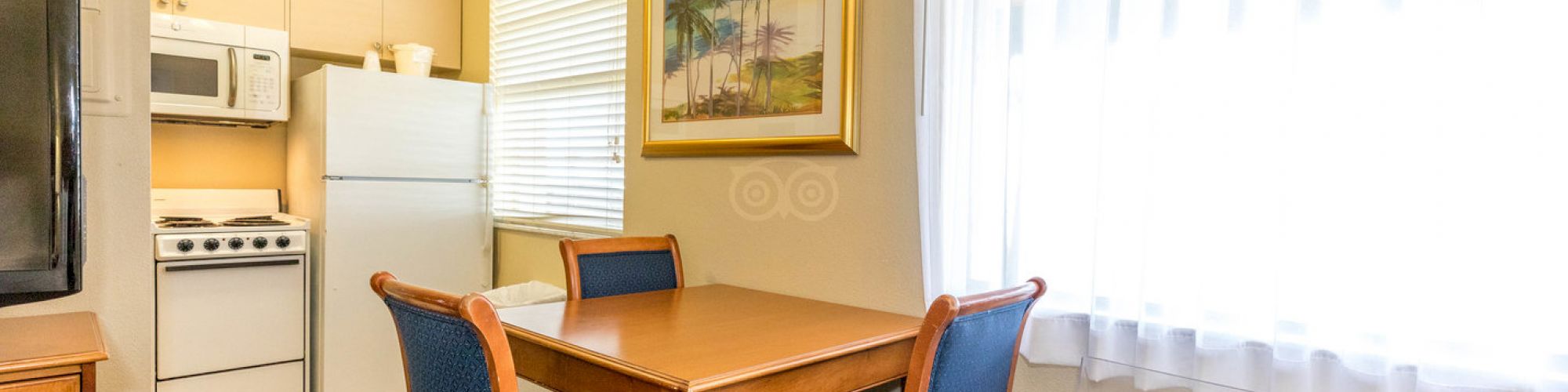 The image shows a small kitchen with a dining table and four chairs, a refrigerator, a stove, a microwave, and a painting on the wall.