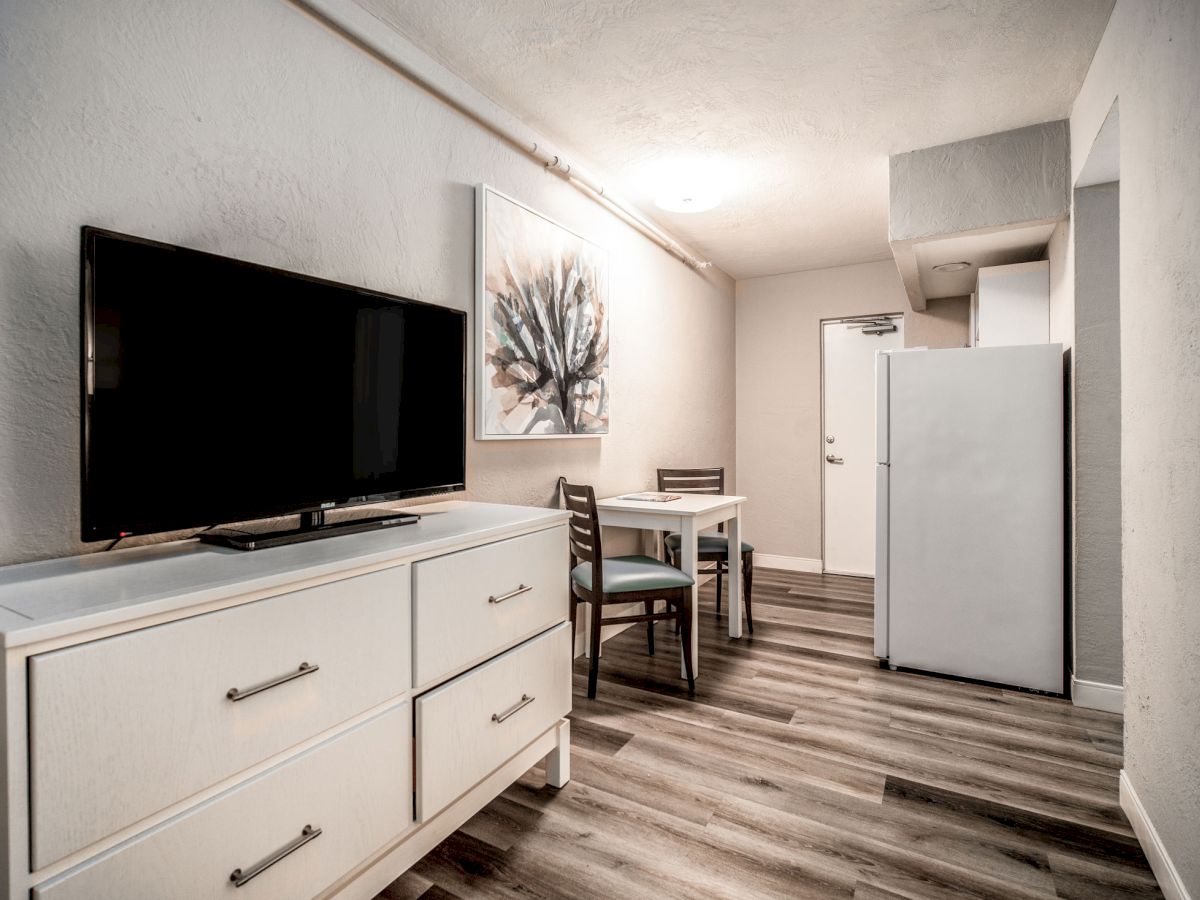 A room with a TV on a drawer unit, a small dining table with chairs, a refrigerator, light flooring, and a wall painting.