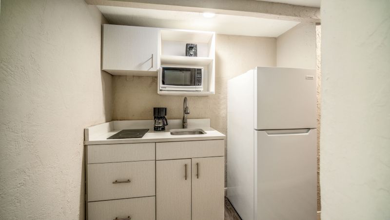 A compact kitchenette with white cabinets, microwave, coffee maker, sink, electric stovetop, and refrigerator.