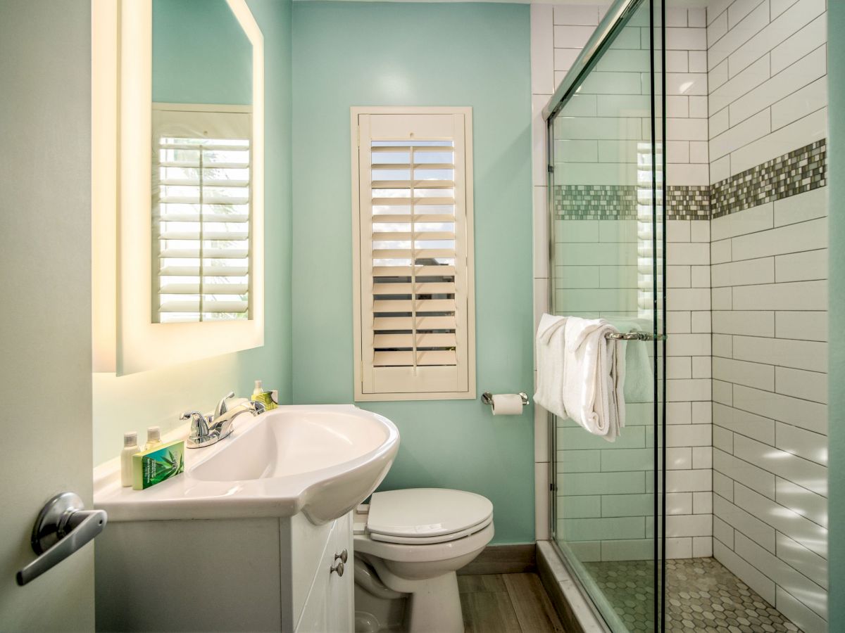 A modern bathroom with a vanity, toilet, and walk-in shower with glass doors, white tiles, and a window with shutters.