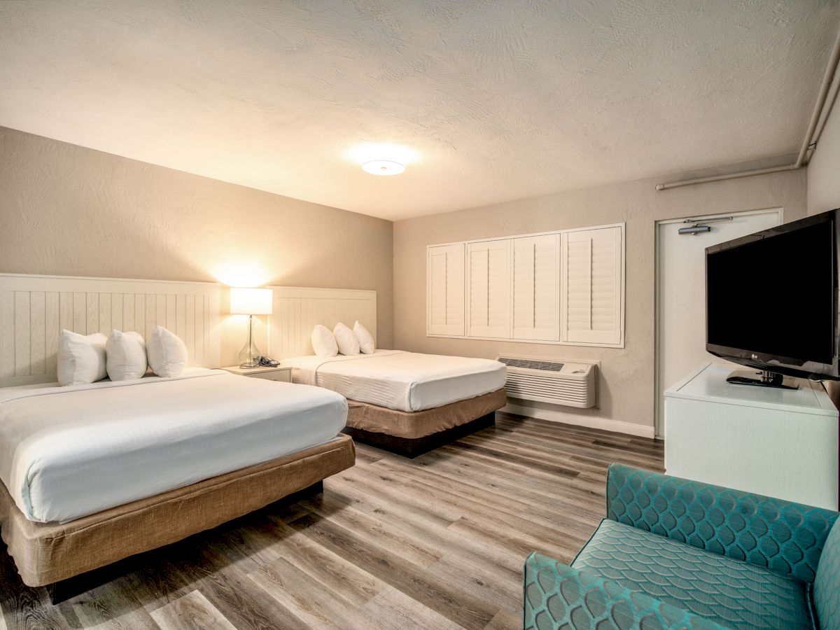 The image shows a hotel room with two beds, a TV, a teal chair, a bedside lamp, wooden flooring, and minimalistic decor, creating a cozy atmosphere.