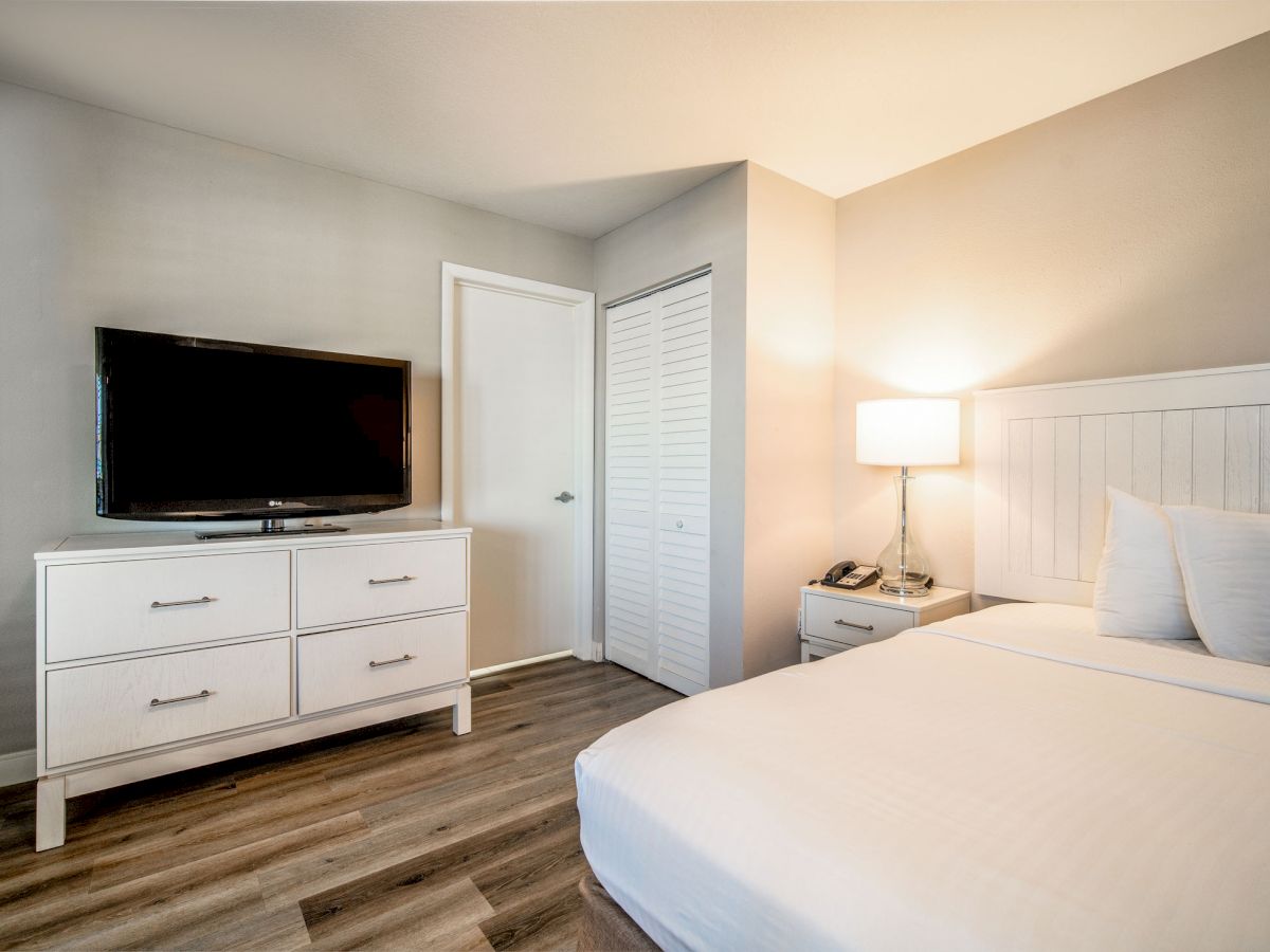 A modern hotel room with a bed, a TV on a dresser, a nightstand with a lamp and phone, and a closet door, featuring light-colored decor.