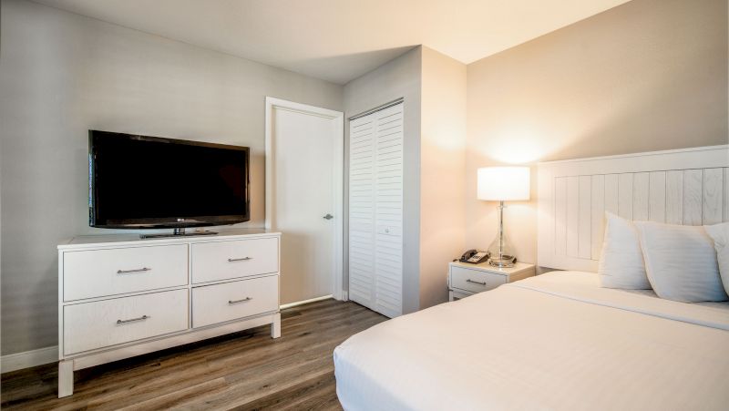 A hotel room with a white bed, dresser with TV, and nightstand with lamp.