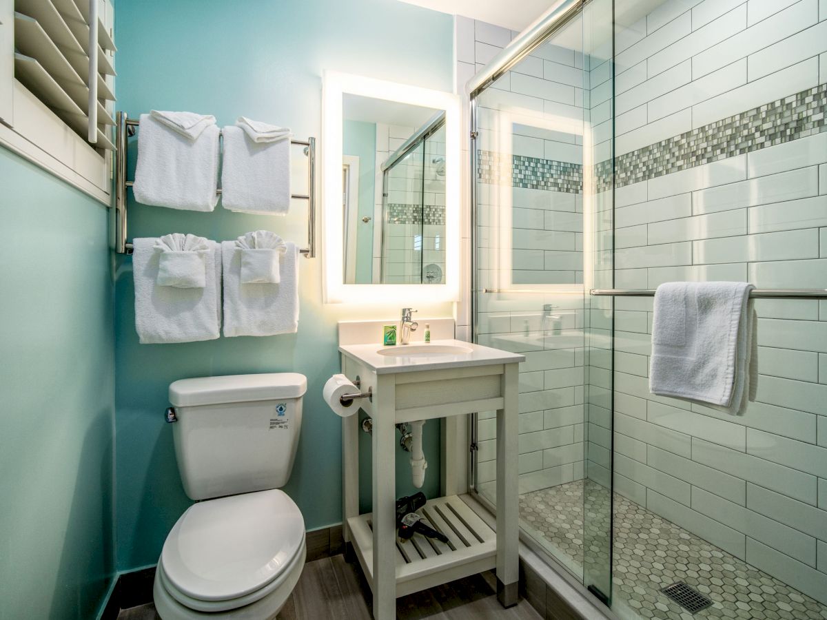 This image shows a modern bathroom with a toilet, a sink with a lighted mirror, towel racks with towels, and a glass-enclosed shower.