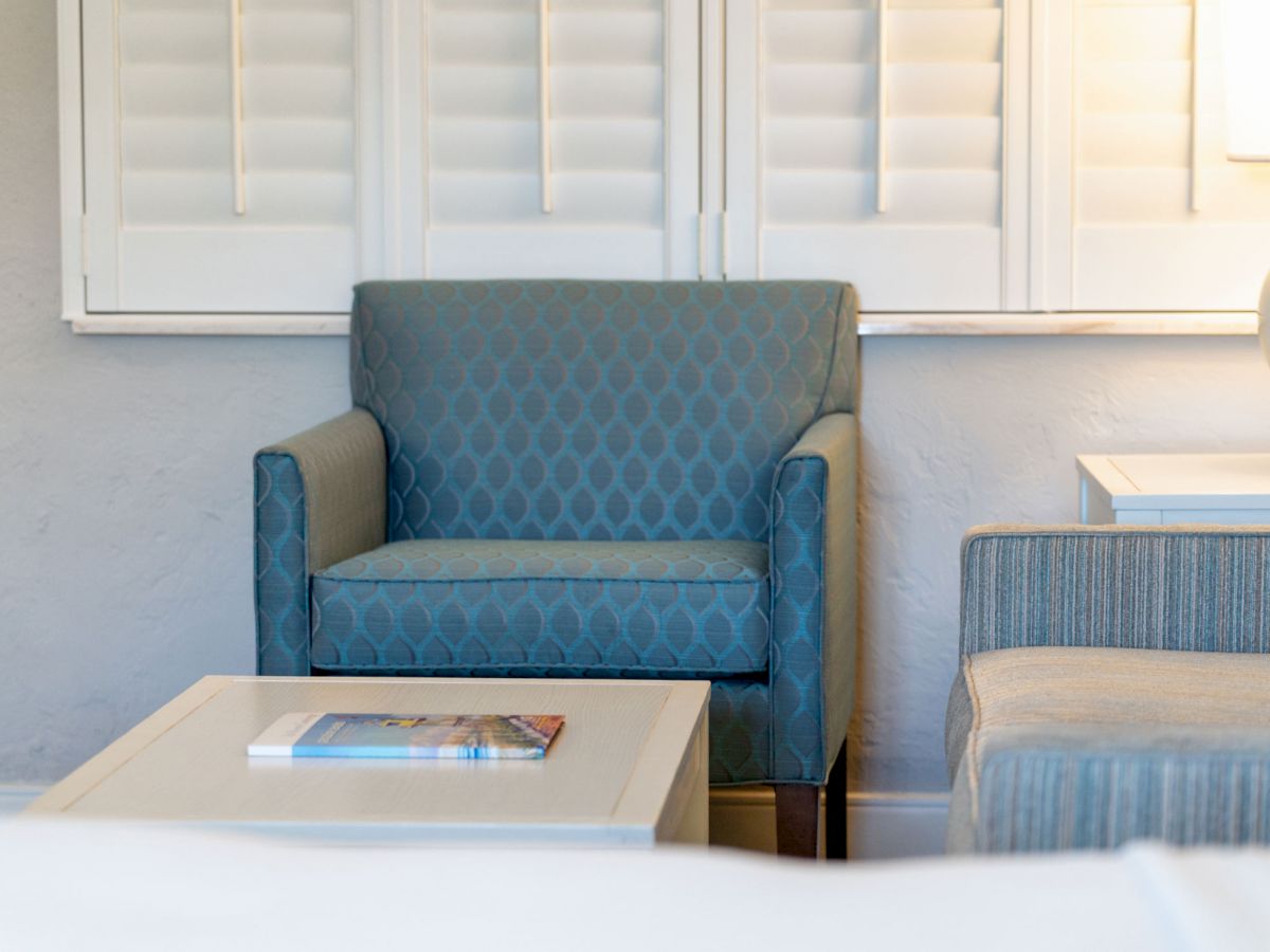 A cozy living space with a blue armchair, white coffee table with a magazine, white shutters, and a lamp on a side table ends the sentence.