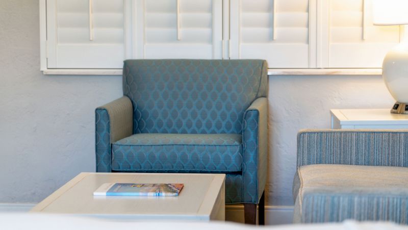 A cozy seating area with a blue armchair, a side chair, a coffee table with a book, and a lamp on a side table.