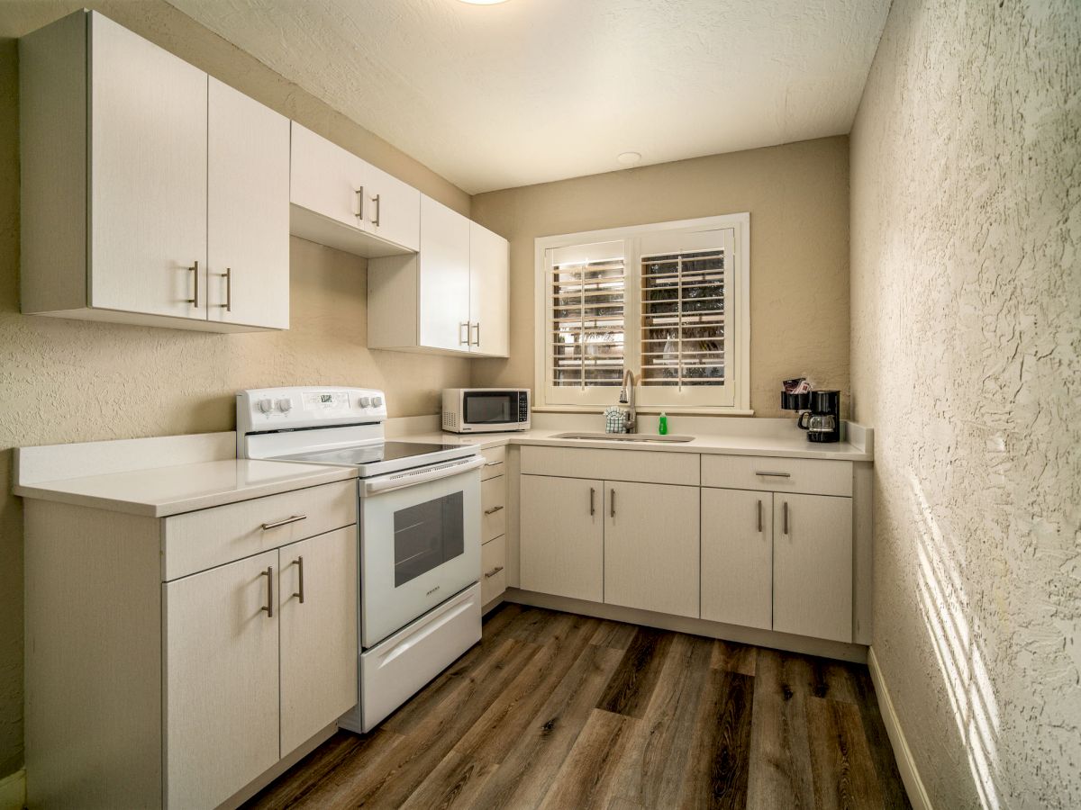 A small kitchen with white cabinets, an oven, microwave, coffee maker, and a window with shutters, set against a textured wall and wooden floor.