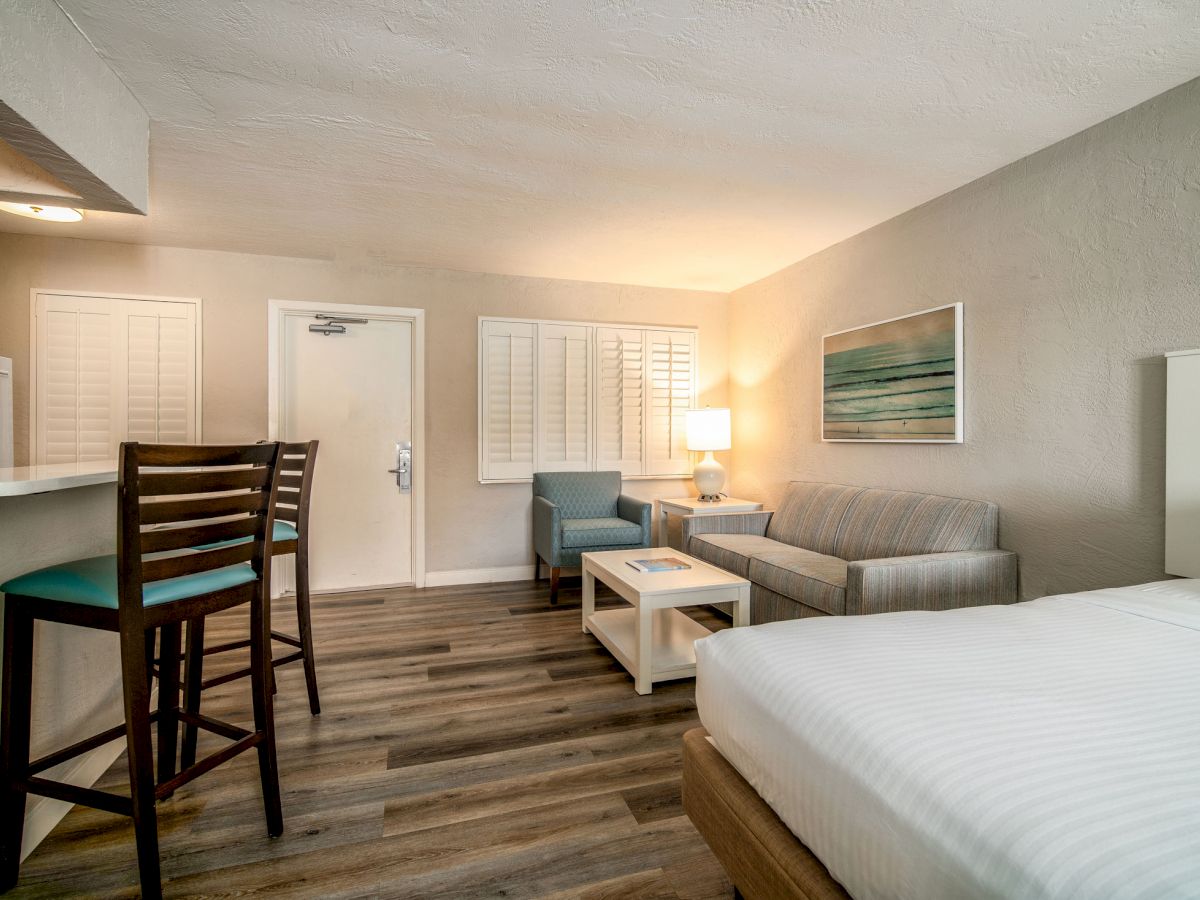 This image shows a modern hotel room with a bed, seating area, bar stools, and a kitchenette. The decor is minimalist with neutral tones.