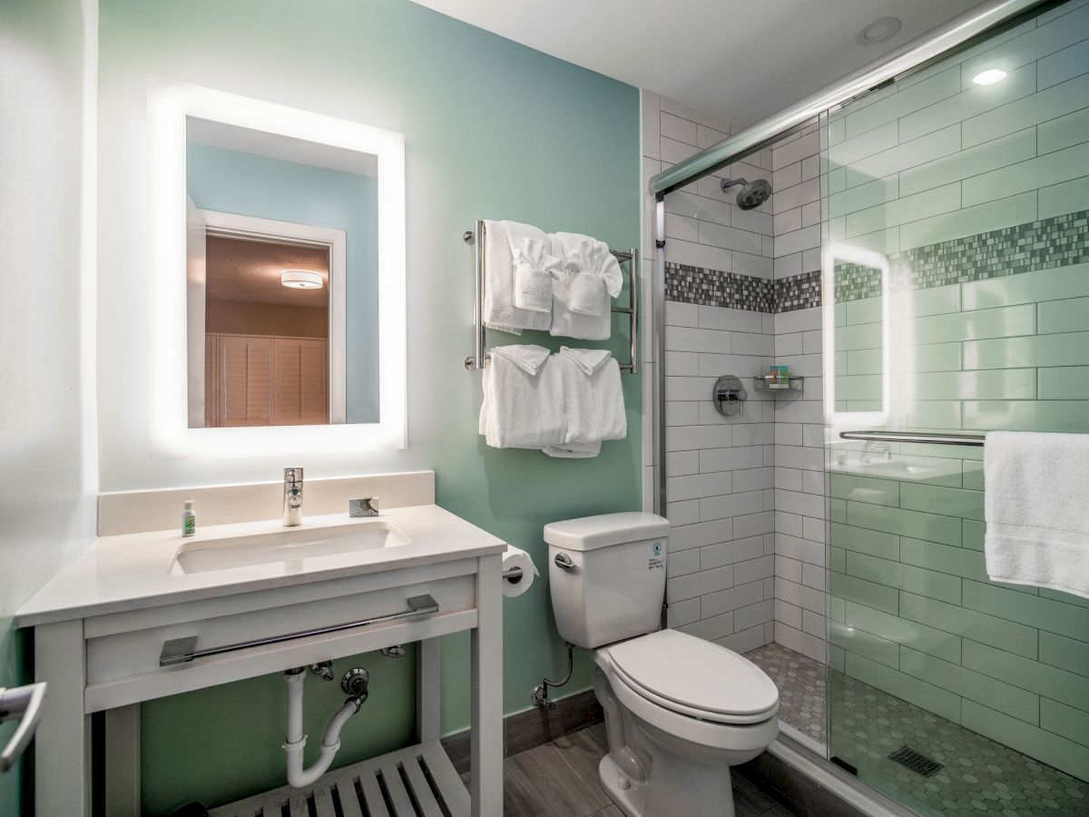 The image shows a modern bathroom with a sink, illuminated mirror, toilet, and shower area. Towels are neatly arranged on a rack above the toilet.