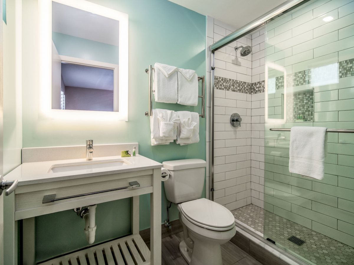 Modern bathroom with a glass-enclosed shower, toilet, wall-mounted sink, and towel racks. White tiles and a backlit mirror add brightness.