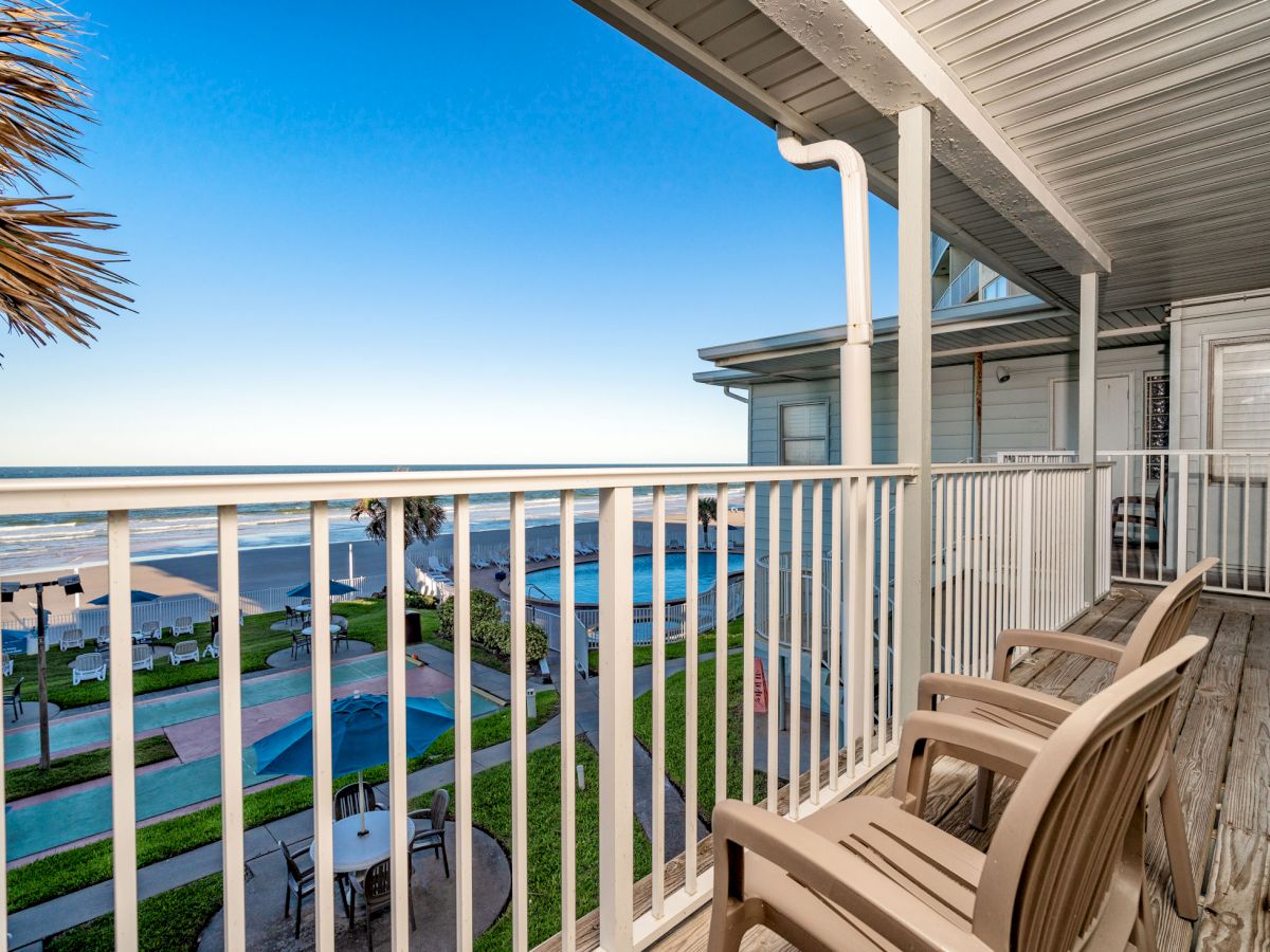 A beachfront balcony with two wooden chairs overlooks a pool area and the ocean, under a shaded cover with clear skies in the background.