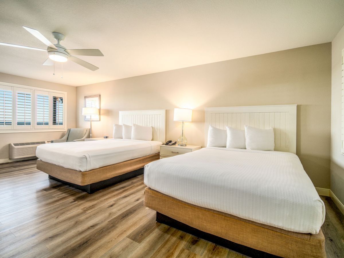 A modern hotel room with two double beds, white bedding, a ceiling fan, wooden flooring, and a window with shutters.