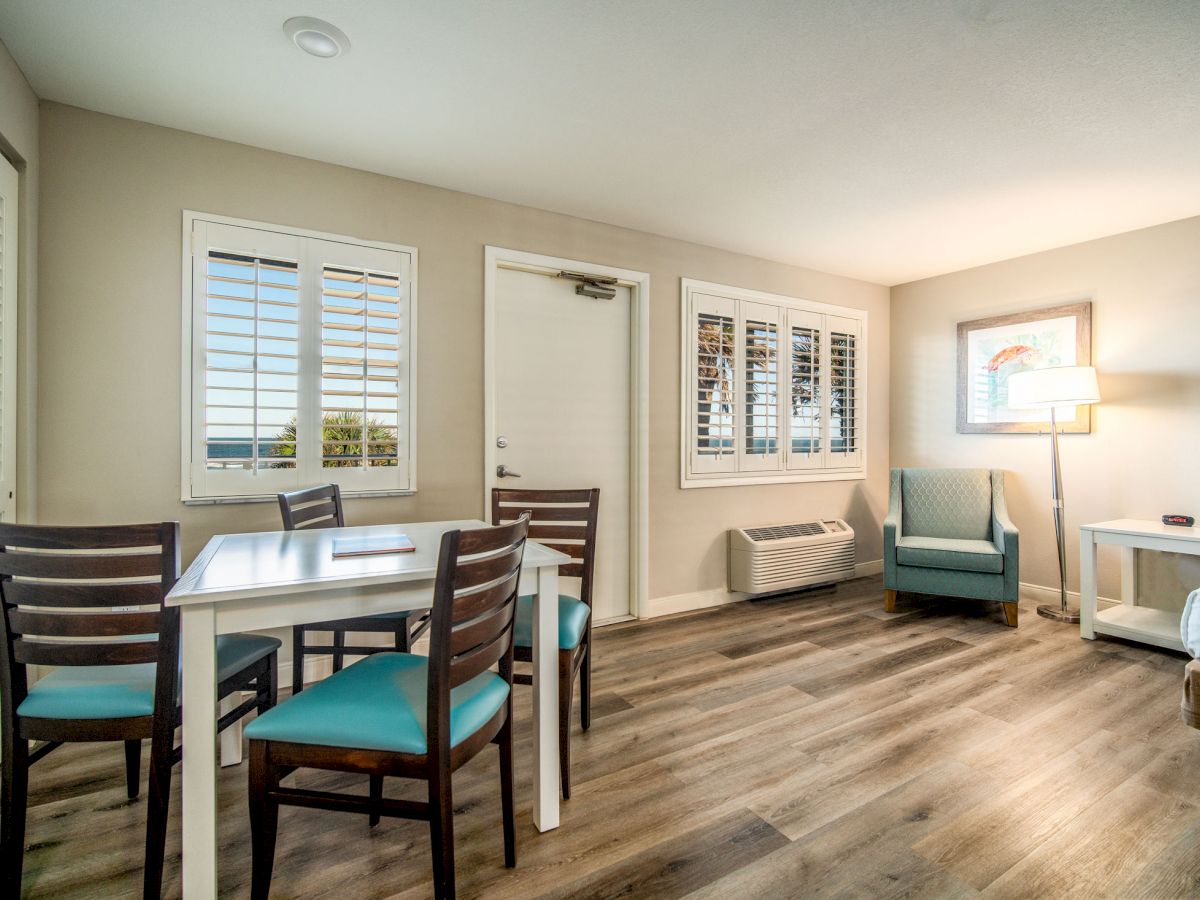 The image shows a modern hotel room featuring a dining table with chairs, an armchair, a standing lamp, and wooden flooring, ending the sentence.