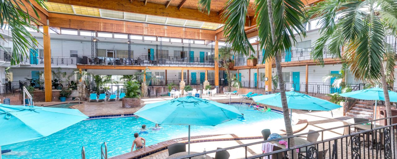 An indoor pool with blue umbrellas and palm trees surrounding it, with people swimming and lounging on the balcony in the background.