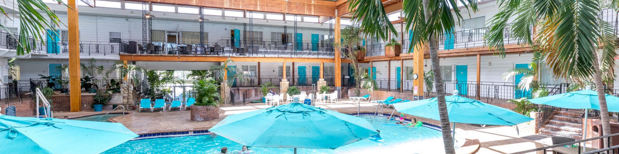 An indoor pool area with blue umbrellas and lounge chairs, surrounded by palm trees and enclosed by a two-story building with balconies, ending the sentence.