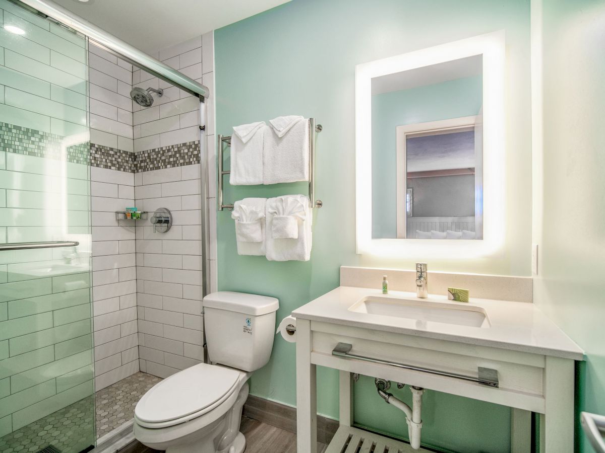 The image shows a modern bathroom with a glass shower enclosure, white sink, illuminated mirror, and towels neatly hung.