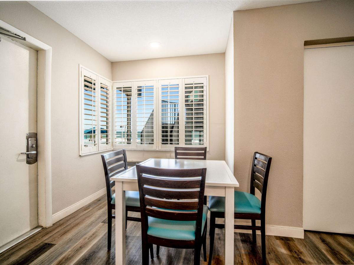 A small dining area with a table and four chairs, next to windows with shutters, hardwood flooring, and beige walls.