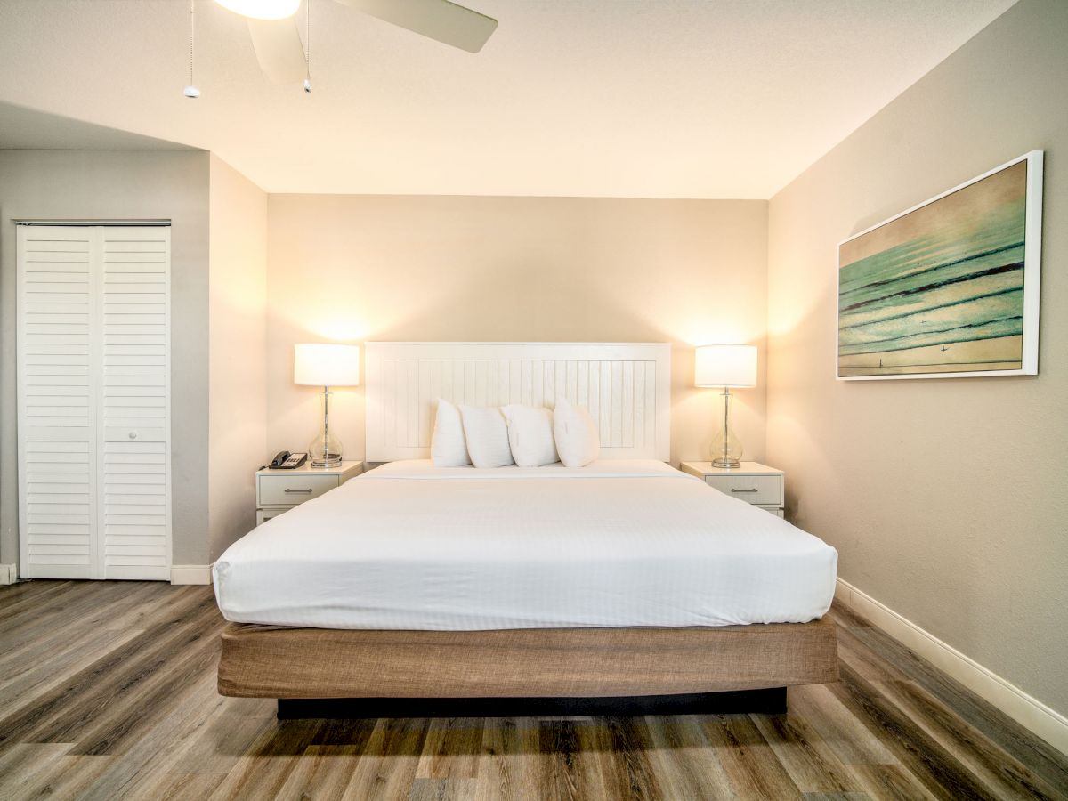 A modern bedroom with a white bed, two lamps on bedside tables, a painting on the wall, wooden flooring, and a ceiling fan.