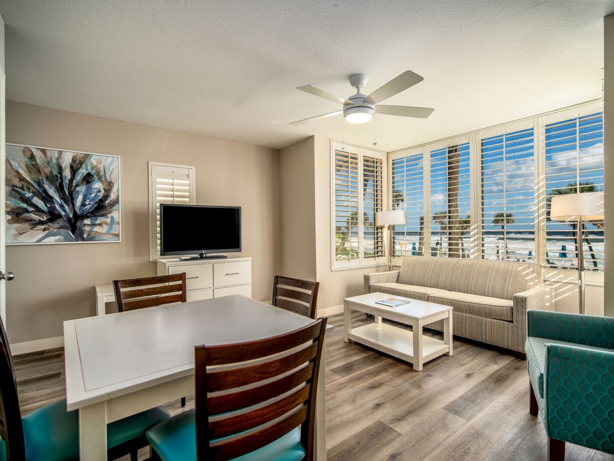 A modern living room with a dining table, chairs, sofa, TV, artwork on the wall, ceiling fan, and large windows offering a scenic view outside.