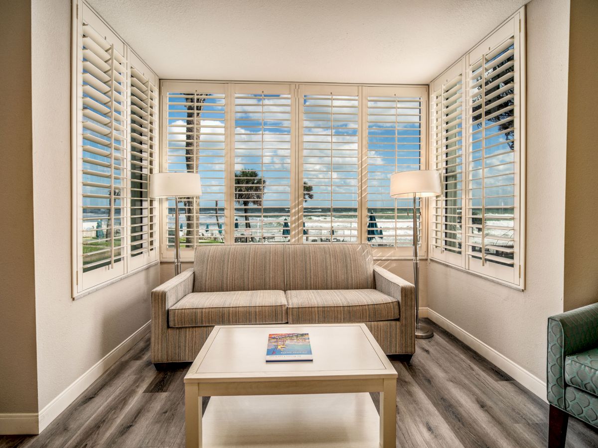 A cozy sitting area with a sofa, coffee table, and armchair, featuring large windows with a beach view and two floor lamps next to the sofa.