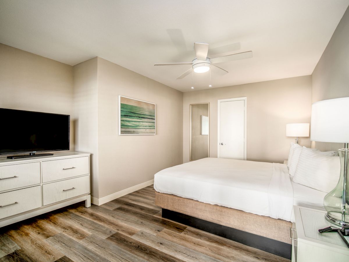 A modern, tidy bedroom with a white bed, television on a dresser, wall art, and a ceiling fan, showcasing a minimalist design.