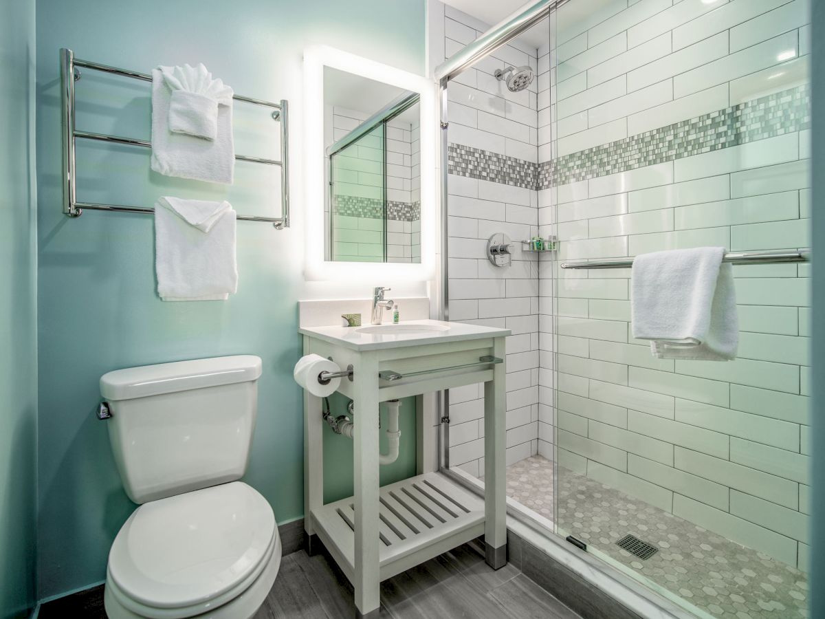 The image shows a modern bathroom with a toilet, vanity with a mirror, towel rack, and a glass-enclosed shower with white subway tiles.