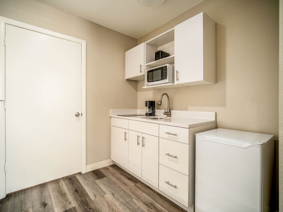 A small kitchenette with white cabinets, a sink, a coffee maker, a microwave, and a mini-fridge beside a closed door on a wood floor.