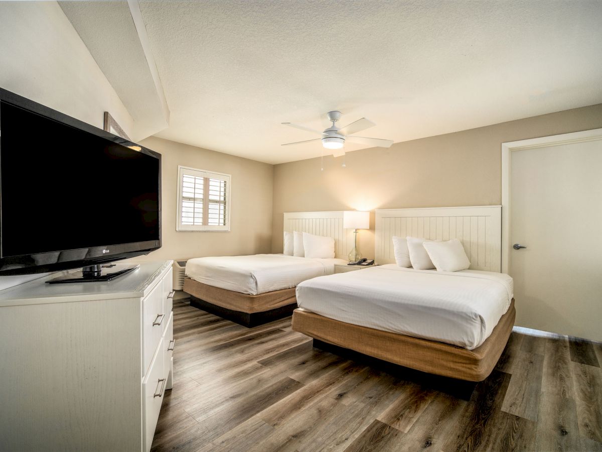 The image shows a hotel room with two beds, a ceiling fan, a flat-screen TV on a dresser, and hardwood floors, creating a clean, modern look.