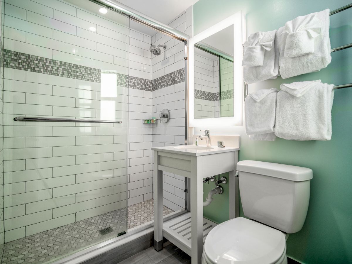 A modern bathroom features a glass-enclosed shower, white brick-patterned tiles, a sink with a mirror, a toilet, and towel racks with neatly hung towels.