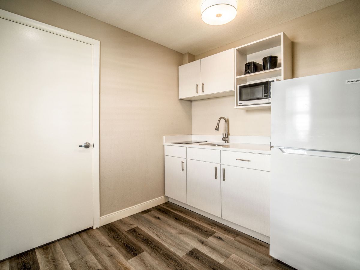 A small kitchenette with white cabinets, a sink, a microwave, and a refrigerator, all set against a wooden floor and light-colored walls.