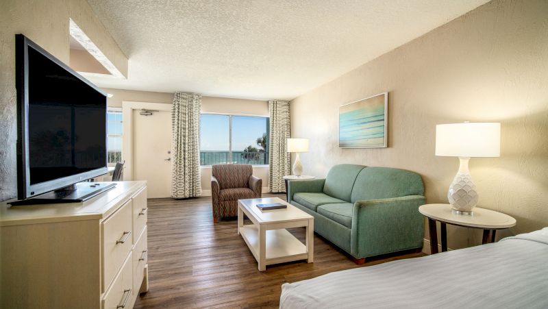 A wide view of a hotel room with a sofa, armchair, coffee table, TV, and bed.