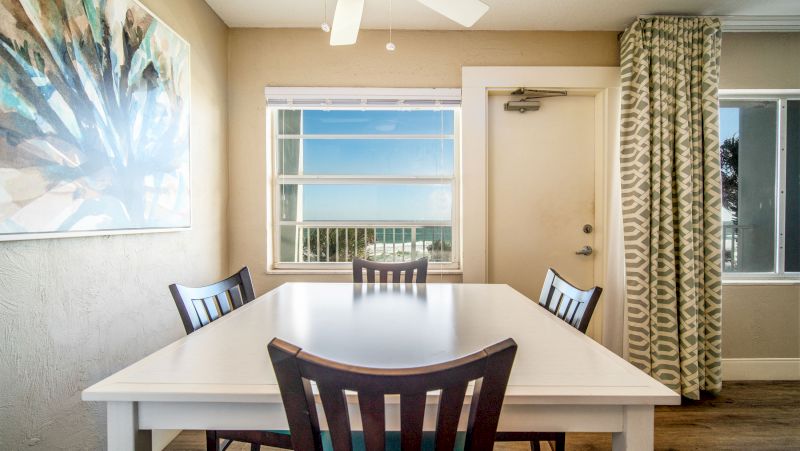 A dining area with a white table and four dark wood chairs, a large window with a view, and a door.