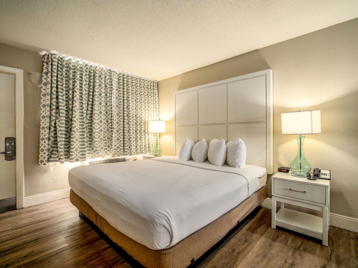 A hotel room features a bed with white linens, nightstands, lamps, patterned curtains, and wooden flooring.