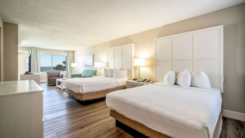 A wide view of a hotel room with two beds, a desk, a window, an armchair, and a coffee table.