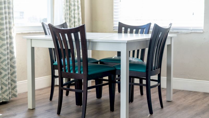 A white dining table with four black chairs and green cushions in a room with beige walls and two windows.