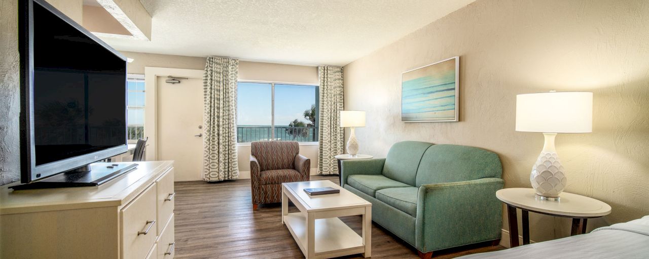 A wide view of a hotel room's living area with a sofa, coffee table, TV, and window.