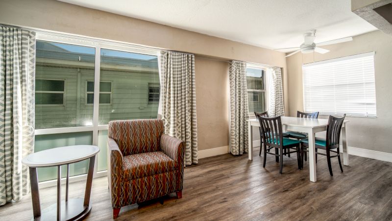 A wide view of a dining area with a table and chairs, an armchair, and large windows with curtains.