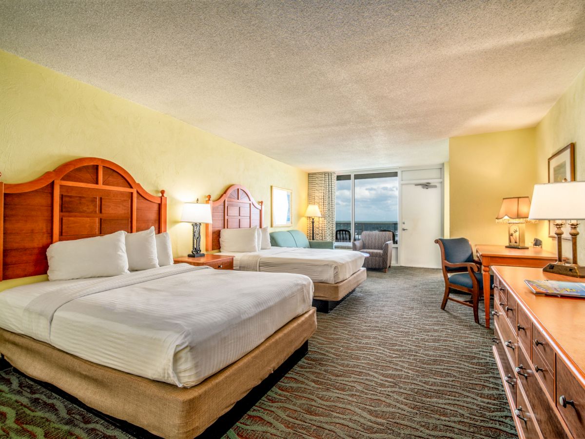 A hotel room with two beds, a desk, a dresser, and lamps, featuring a window with a view.
