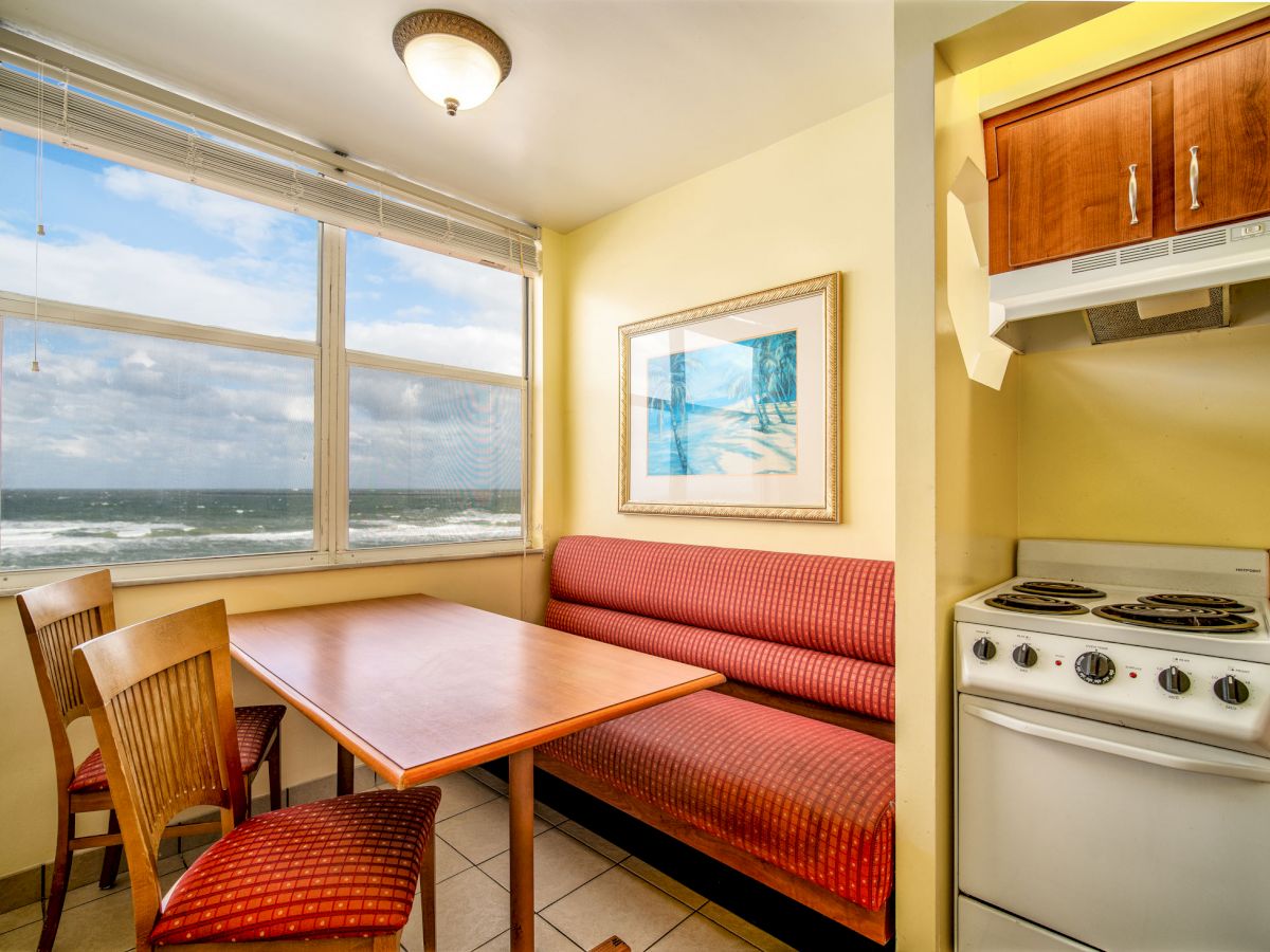 The image shows a small dining area with a table, chairs, and a red bench, next to a window with an ocean view. A kitchenette is on the right.