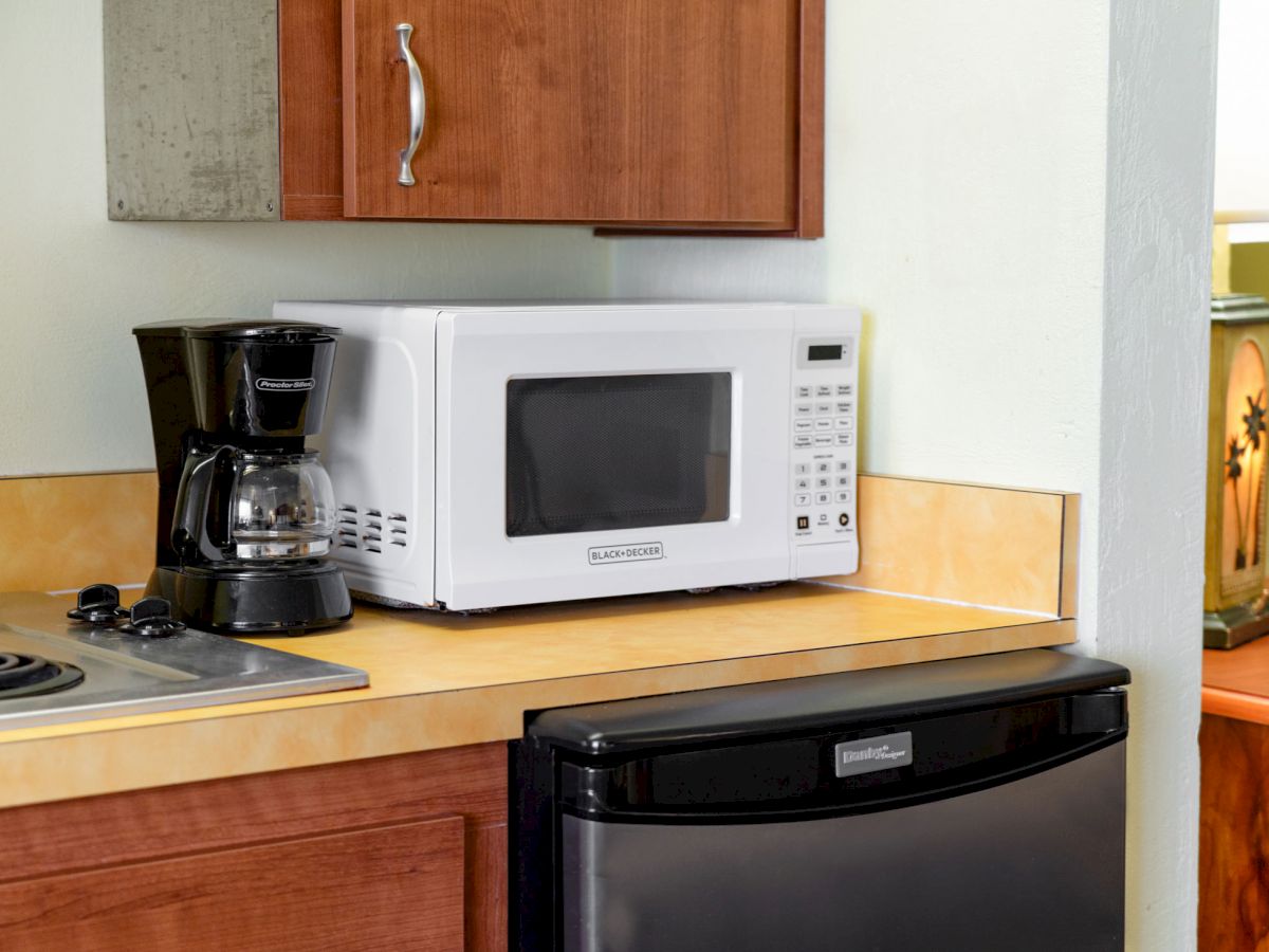 A kitchen countertop with a coffee maker, microwave, and mini fridge beneath, next to a stovetop with a wooden cabinet above.