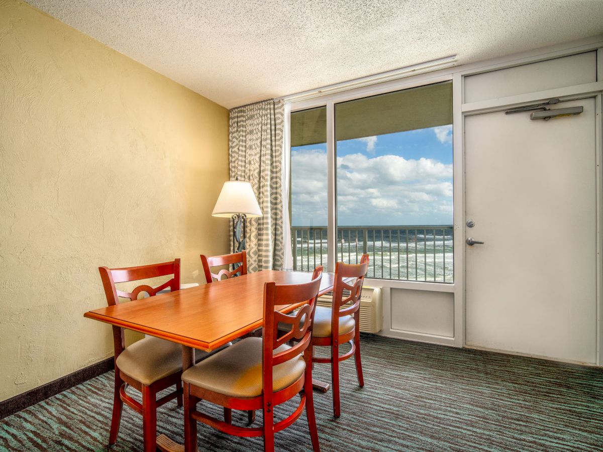 A dining area with a wooden table and four chairs, a lamp on the left, large window with ocean view, and a door next to the window, ending the sentence.