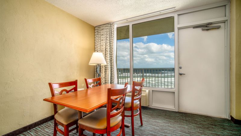 A hotel room dining area with a table, chairs, and a lamp, overlooking the beach through a large window.
