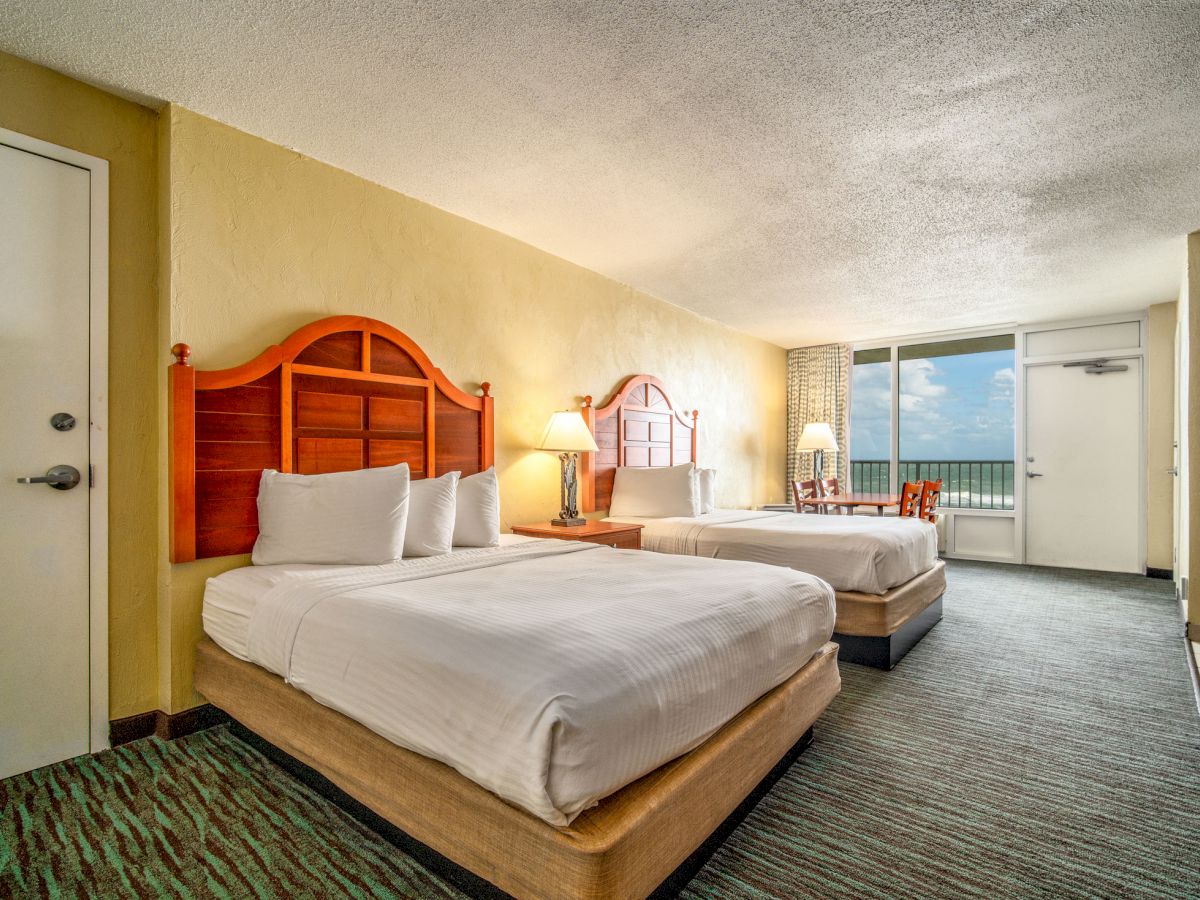 This image shows a hotel room with two double beds, a small table with chairs near a window, and a door leading to a balcony or outside view.